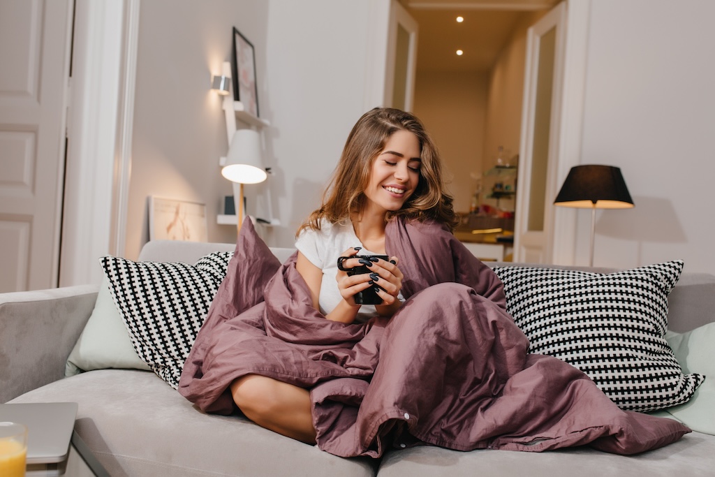 Cheerful girl sitting on couch with blanket and cushions and smiling. Spectacular brunette lady laughing, while drinking coffee under plaid..